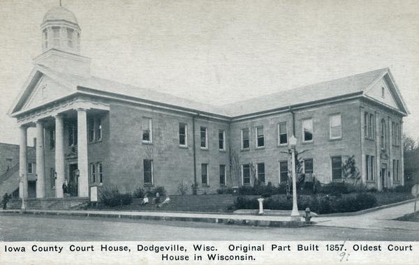 Iowa County court house with people sitting on the lawn. The wing on the right was added in 1927. Caption reads: "Iowa County Court House, Dodgeville, Wisc. Original Park Built 1857 [or 1859]. Oldest Court House in Wisconsin."