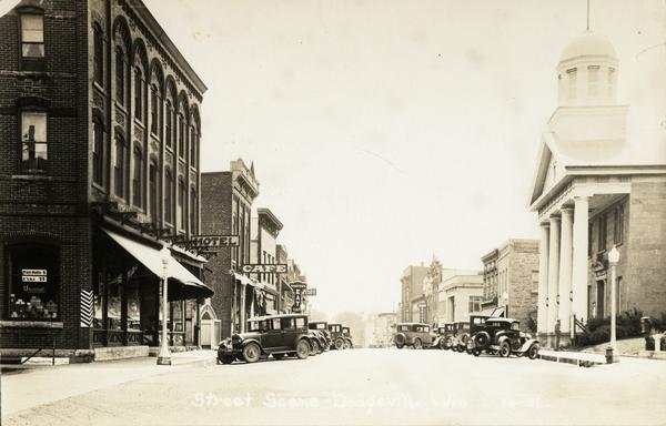 Main street, with Iowa County Courthouse on the right, and a hotel and cafe on the left. Parked cars line the street. Caption reads: "Street Scene — Dodgeville, Wis."