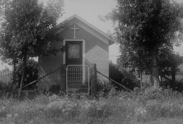 One-room building out in a field, with a cross above the doorway.  Trees stand to the left and right of the shrine, and a fence and gate surround it.