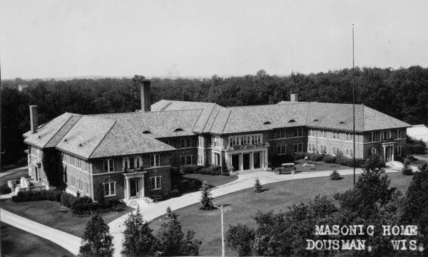 Elevated view of the Masonic Home, surrounded by a lawn and trees. There are two flagpoles along a driveway leading around the building. Caption reads: "Masonic Home, Dousman, Wis."