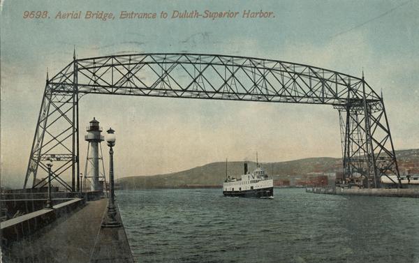 Aerial bridge and harbor, with a large boat passing underneath. A lamppost and a beacon are along the walkway towards the bridge on the left. Caption reads: "Aerial Bridge, Entrance to  Duluth-Superior Harbor."
