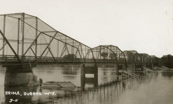 View towards the bridge along the left. Trees are along the opposite shoreline. Caption reads: "Bridge, Durand Wis."