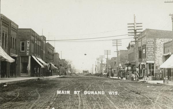 View down unpaved Main Street. Several businesses line the street and large utility poles run the length of it. A sign on the side of a building on the left reads: "Clothing," and a sign on a building on the right reads: "Hardware Stoves Sash Doors Paints New Furniture Store Carpets Trunks Lowest Prices." Caption reads: "Main St., Durand, Wis."