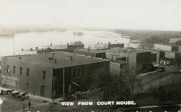Elevated view of commercial buildings along the river from the vantage point of the Court House. Horse-drawn carriages and pedestrians are along the street in the foreground. Caption reads: "View from Court House."