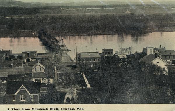 Elevated view of Durand. A street in the foreground leads to a bridge that crosses the river. Caption reads: "A View from Morsbach Bluff, Durand, Wis."