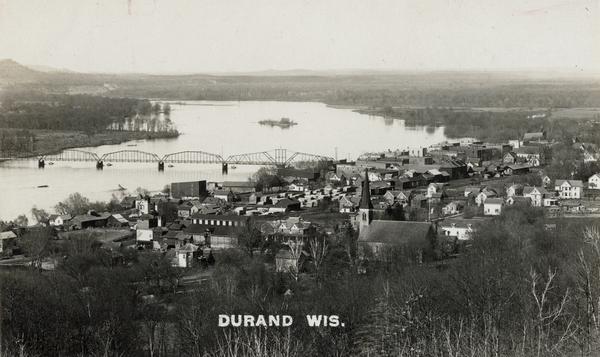 Elevated view of Durand from a high vantage point, looking towards the river. A bridge spans the river, and in the foreground just below the hill is a church with a tall steeple. Caption reads: "Durand Wis."