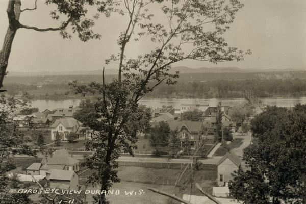 Elevated view from hill of Durand through trees, with houses in the foreground, and the river and hills in the background. Taken in the summer, with wash on clotheslines. Caption reads: "Bird's Eye View of Durand, Wis."