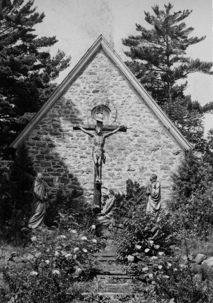 Statues depicting the crucifixion of Christ, surrounded by flowers before the chapel. Two pine trees are behind the chapel.