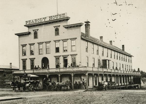 Front view of the inn. Several men and women are either standing or sitting on the balcony, and several people are standing on the porch under the balcony. Three horse-drawn wagons and carriages are at the front and side of the building.