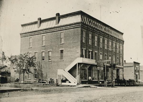 Three or four-story brick building, with a "Drug Book Store" sign at the front, and a stair wrapping around the side of the building up to a balcony. A man is standing on the sidewalk in front of the building under the balcony, and two horse-drawn wagons are parked along the curb.