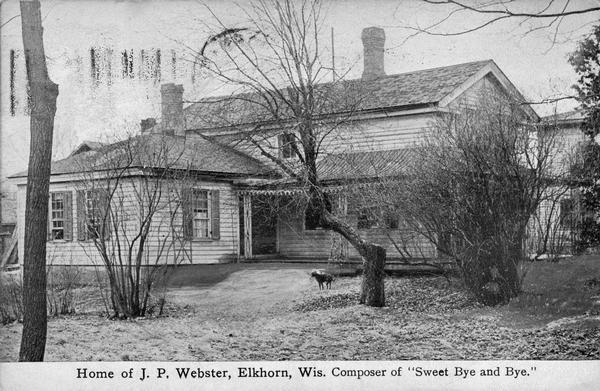 Front view of the home of composer Joseph Philbrick Webster, with trees and a small dog in the yard. Joseph Philbrick Webster composed "Sweet By and By" and the Civil War favorite, "Lorena". He lived in the house from 1857 until his death in 1875 and most of his more than 1000 songs were written there. Built in 1836 as a federal land grant office, the structure now serves as a museum. Caption reads: "Home of J. P. Webster, Elkhorn, Wis. Composer of 'Sweet Bye and Bye."