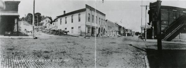 Panoramic view looking down Main Street, with horses and carriages parked outside the storefronts on the street.