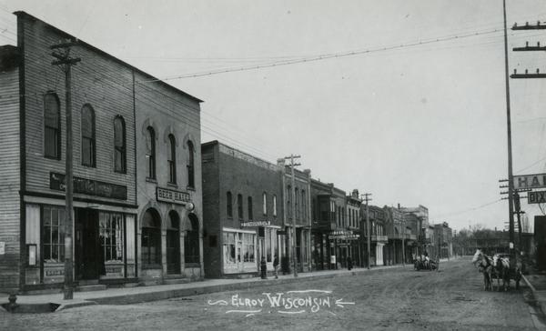 View across Main Street towards the left side of the street. Horse-drawn vehicles are along the curbs. Caption reads: "Elroy, Wisconsin."