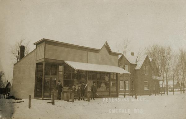 A group of men with children are standing outside a storefront on the Peterson Block in Emerald. Caption reads: "Petersons Block, Emerald Wis".