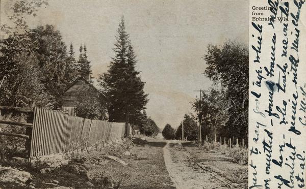 Fence along road, with house in background. Caption reads: "Greetings from Ephraim, Wis."