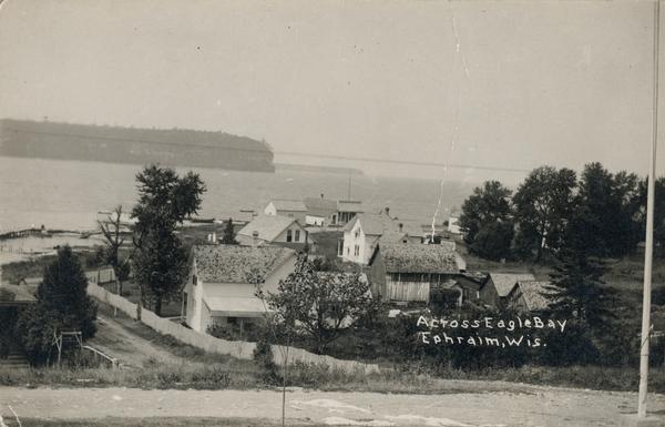 View from hill across Eagle Bay with buildings and trees in the foreground. Caption reads: "Across Eagle Bay, Ephraim, Wis."