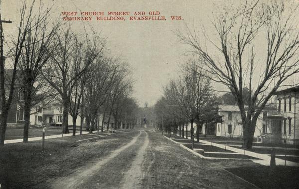 Looking down unpaved West Church Street, lined with trees. Caption reads: "West Church Street and Old Seminary Building, Evansville, Wis."