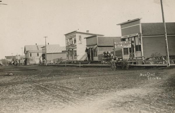 View across unpaved Main Street, with storefronts on the right, including a restaurant and meat market. Caption reads: "Main Street".
