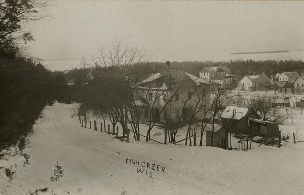 View from hill towards downtown Fish Creek, with the buildings and ground covered in snow. There is a road in the foreground leading off to the left. The bay is in the distance. Caption reads: "Fish Creek Wis."