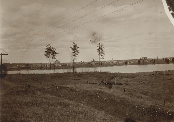 Field and trees and water are in the foreground. On the far shoreline are buildings in the town.