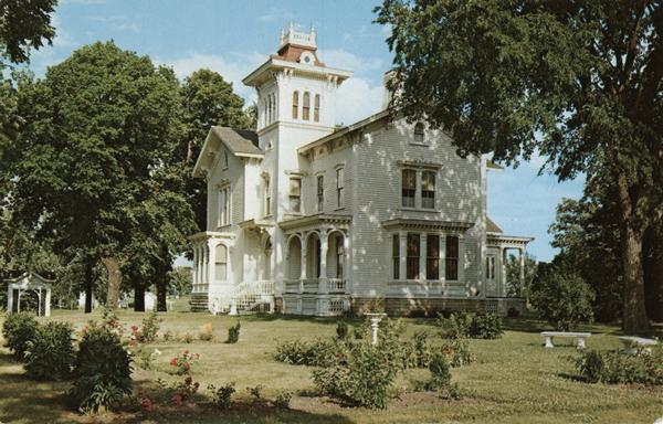 Galloway House Museum and flower-laden lawn on a sunny, summer day.  Large oak trees stand to the left and right of the museum.  Two benches, a bird bath, and a gazebo or an arbor are on the lawn.