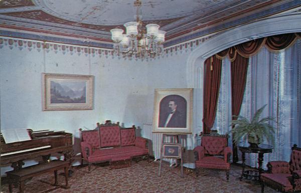 Interior view of the parlor, with a piano on the left, a sofa to the right in the corner of the room, and a portrait on an easel in the center. There is a plant on a table on the right between two chairs i front of an archway framing windows with drapes. A landscape painting hangs on the wall, and a chandelier hangs from the intricately-decorated ceiling.