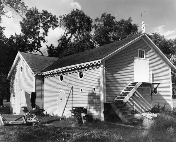 View of the exterior of the carriage house in the spring or summer, with miscellaneous tools and other items littered throughout the yard. A wooden stairway is at the back right wall of the carriage house. The house has semi-circular eaves running along the side of the building.