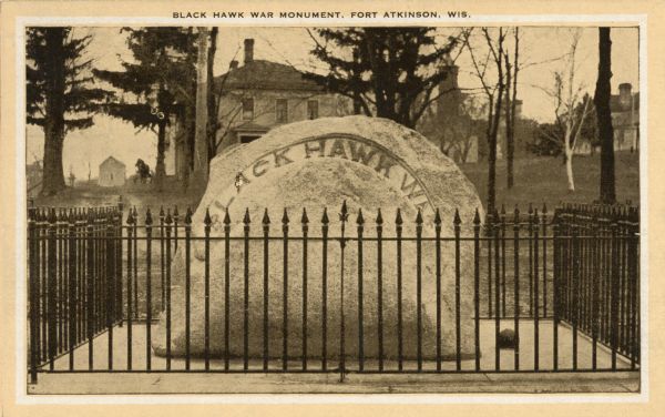 Black Hawk war monument behind a fence, located at Fort Atkinson. Buildings are in the background. Caption reads: "Black Hawk War Monument, Fort Atkinson, Wis."