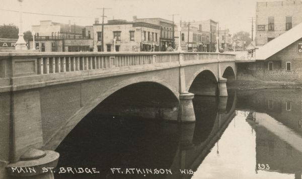 View of a river and Main Street bridge above the river. Commercial buildings line the street on the opposite side of the bridge. Caption reads: "Main St. Bridge, Ft. Atkinson, Wis."