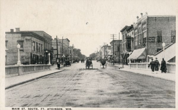 View down center of bridge looking down Main Street, with horse-drawn carriages and people on the sidewalk. Caption reads: "Main St. South, Ft. Atkinson, Wis."