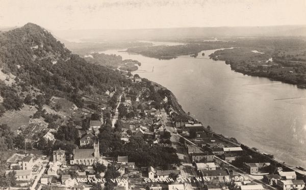 Bird's-eye view of Fountain City looking south, with high bluff on the left, and the Mississippi River on the right. Fountain City, about 4 blocks wide, nestles between the bluff and the river. Streets are unpaved, sidewalks appear to be paved or of wood. Single track railroad line runs along the river, with a steam-powered train at the bottom right corner headed toward the center. Caption at bottom reads: "Aeroplane View, Fountain City, Wis."