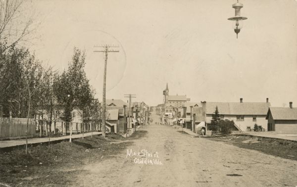 View of Main Street when it was a dirt road. A church is in the background, and telephone poles are along the left side of the road. A streetlight is hanging above the road in the right foreground. Caption reads: "Main Street, Glidden, Wis."