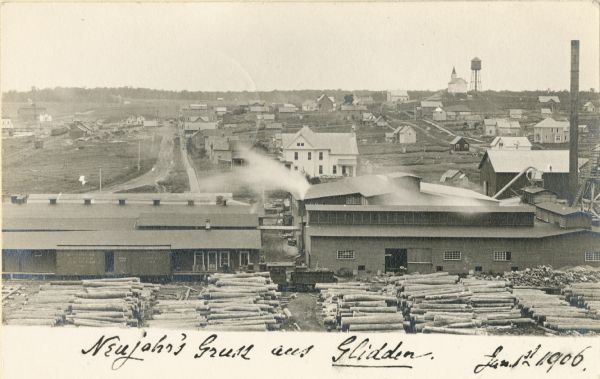 Elevated view of a sawmill and the distant town in the background. Stacks of logs are in the lumberyard. Along the bottom is a New Year's greeting in German.