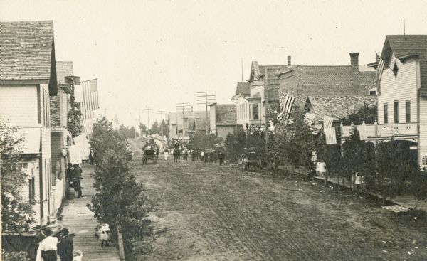 Slightly elevated view looking down Main Street, with people walking on the wooden sidewalks, horse and carriages driving down the street and storefronts with U.S. Flags hanging from windowsills.