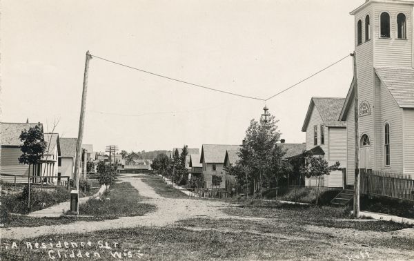 View down road, with homes in a residential area, and a church building in the foreground on the right. The sign above the church door reads: "E. V. Luth. Dreieinigkeits 1888 Kirche". A sign on a building in the distance reads, in part: "[undreadable] Opera House".