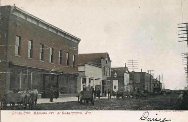 Caption reads: "South Side, Madison Ave. at Grantsburg, Wis." View across road towards pedestrians and horse-drawn wagons at the curb.