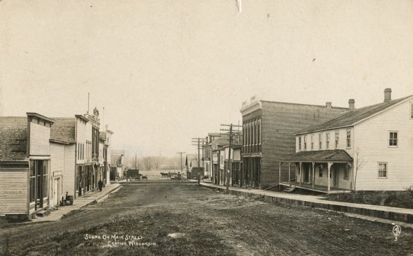 View down unpaved Main Street with storefronts are along both sides. A sign on a building further down on the right reads: "Nixon House". Caption reads: "Scene on Main Street, Gratiot, Wisconsin."