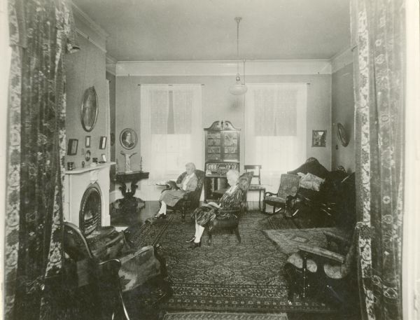 Interior of the front parlor, with two women sitting on rocking chairs in front of the fireplace. The house is located at 203 South Jefferson Street. Beaumont's house was demolished in 1932.