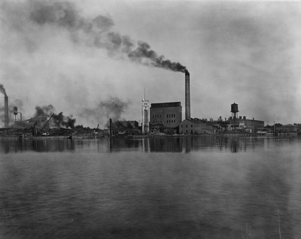 Northern Paper Mills' chimney, water towers, buildings, and lumber yard, seen from the Fox River.