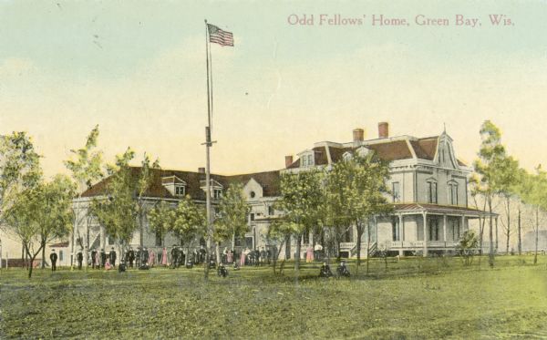 A large gathering of men and women stand outside the Odd Fellow's Home. There is a flagpole on the grounds with an American Flag among trees. People are posing on the lawn and near the building. Caption reads: "Odd Fellows' Home, Green Bay, Wis."