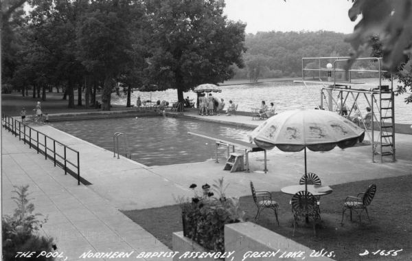View towards swimming pool towards people relaxing and sitting around the pool near the lake at the Baptist Assembly Grounds. Caption reads: "The Pool Northern Baptist Assembly, Green Lake, Wis."