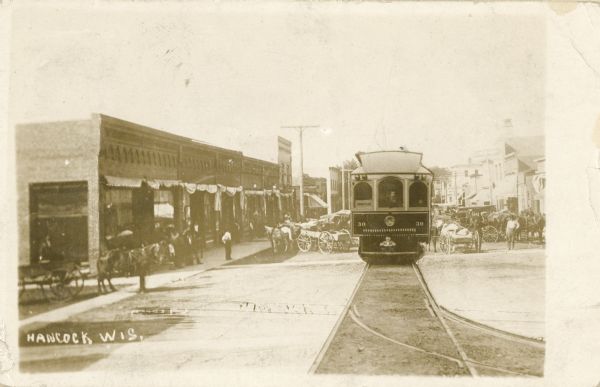 View of downtown Hancock with a streetcar driving down the street and people walking on the sidewalk and crossing the street. A streetcar has been superimposed into the image. Caption reads: "Hancock Wis."
