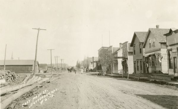 View down unpaved East Main Street, with lumber stacked along the roadside on the left, and on the right storefronts are lining the street. A horse-drawn vehicle and pedestrians are on the street in the background.