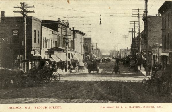 Business district on Second Street lined with horse-drawn vehicles. Caption reads: "Hudson, Wis. Second Street."