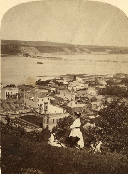 View of Hudson with three women on a hill in the foreground, and the town along the St. Croix River below in the background.