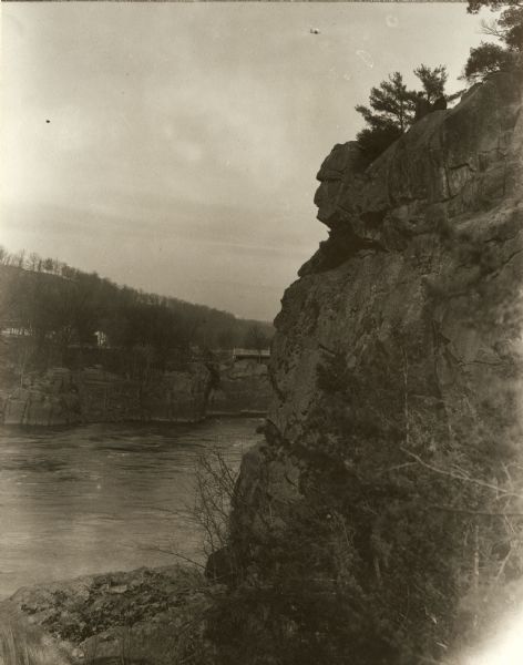 View of the rock formation known as the "Old Man of the Dalles" in the Interstate State Park near Saint Croix Falls, Wisconsin. The St. Croix River is in the background with wooded landscape and a few farm buildings. The St. Croix River bridge connecting Wisconsin and Minnesota is visible at left center.