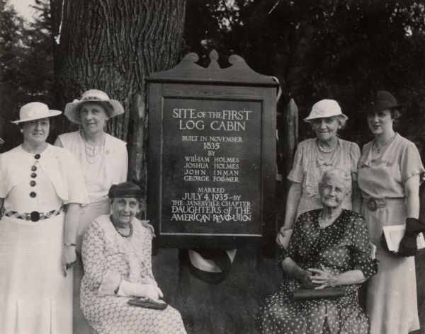 A marker for the first log cabin built in the Janesville area, surrounded by several women. The cabin was built in 1835 by William Holmes, Joshua Holmes, John Inman and George Follmer. The marker was erected by the local Daughters of the American Revolution on July 4, 1935.