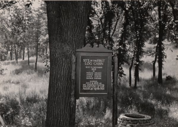 A marker for the first log cabin built in the Janesville area. The cabin was built in 1835 by William Holmes, Joshua Holmes, John Inman and George Follmer. The marker was erected by the local Daughters of the American Revolution on July 4, 1935.