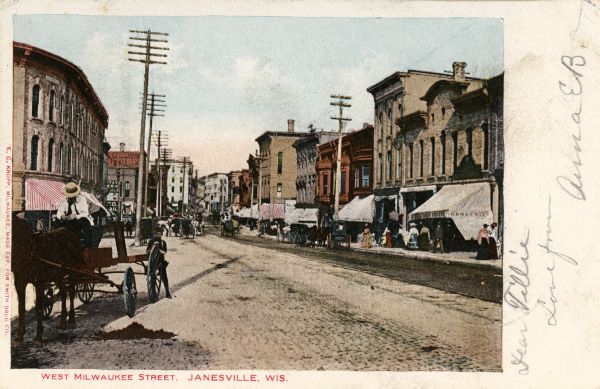 View down left side of street towards the right side of the street. A man is on a horse-drawn wagon in the left foreground. Storefronts are on the right. Caption reads: "West Milwaukee Street, Janesville, Wis." 