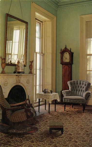 View of chairs and a table set around the fireplace. A mirror is above the fireplace, and a grandmother clock is against the far wall. The windows flanking the corner of the room are at floor height.